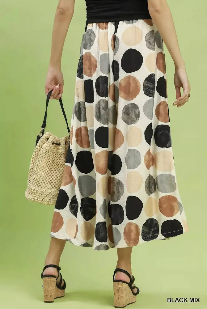 A model from the waist down wears black wide leg pants with a large abstract polka dot pattern in black, gray, beige, and brown on a cream background. She is holding a woven straw bucket bag and wearing cork wedge sandals, set against a solid light green studio background.