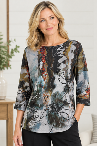 A woman wearing a Ali Miles three-quarter sleeve top with an abstract painterly print in black, gray, white, blue, and rust orange, paired with black pants, standing in a bright indoor setting.