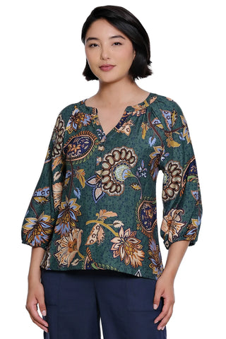 A woman modeling a dark green V-neck blouse with a multicolor floral and paisley print, paired with navy blue pants, against a plain white background.