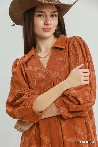 A woman wearing an Umgee terracotta rust-colored long-sleeve collared shirt dress with a tonal leaf pattern, styled with a brown cowboy hat against a light gray background.