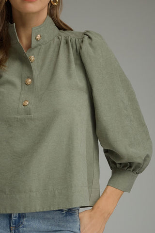 A close-up of a woman wearing a sage green long-sleeve blouse with a high stand collar, gold-tone buttons, and gathered puff sleeves, paired with blue jeans against a neutral gray background.