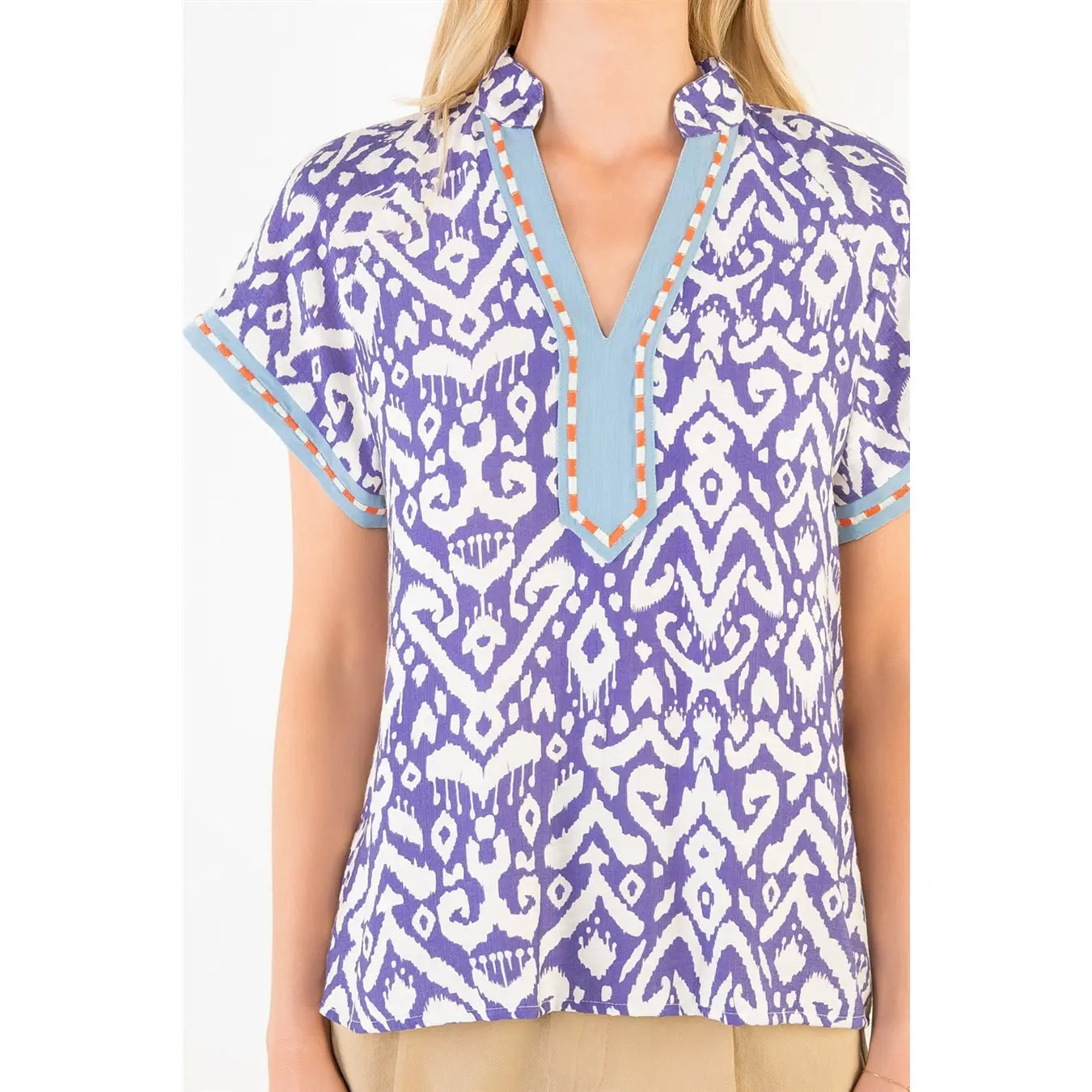 A woman wearing a THML short-sleeve V-neck blouse with a purple and white pattern, light blue trim, and orange stitching details.