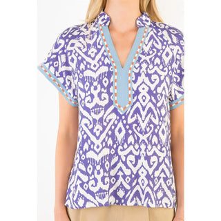 A woman wearing a THML short-sleeve V-neck blouse with a purple and white pattern, light blue trim, and orange stitching details.