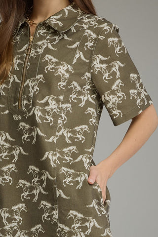 A close-up of a model wearing a short-sleeve collared denim shirt dress in olive green with a white horse pattern, featuring a half-zip neckline.