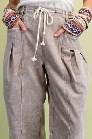 Close-up of a person wearing Easel loose, faded taupe-brown drawstring pants with pleated side details and pockets