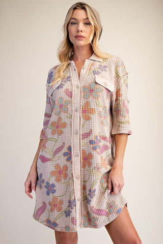 A woman modeling an Easel short button-front shirt dress with a waffle-textured fabric and a pastel floral print in pink, purple, orange, and blue on a light beige base.
