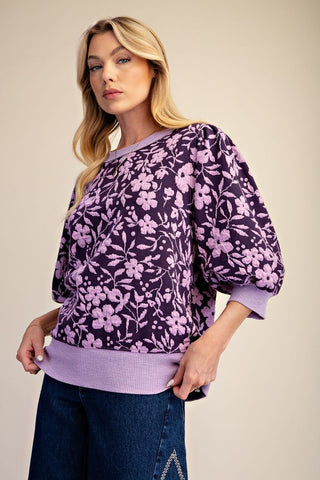 A woman wearing a dark purple and lavender floral print sweatshirt with ribbed trim, paired with dark blue jeans, against a solid beige background.