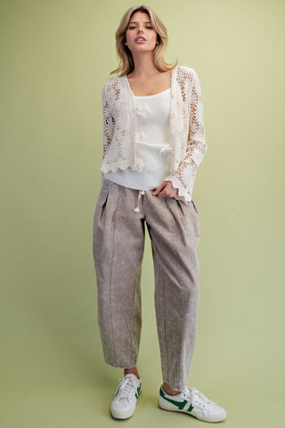 A woman stands wearing a white lace-trim cardigan over a white top, Easel loose taupe drawstring pants, and white sneakers with green accents.