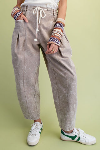 A person wearing Easel loose, faded mushroom colored drawstring pants with pleated details, paired with white sneakers.