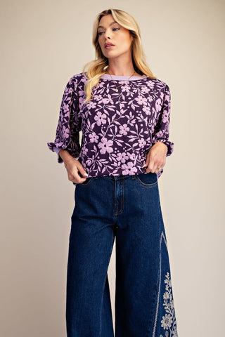 A woman modeling a purple and lavender floral print 3/4 sleeve top paired with dark blue wide-leg jeans, set against a solid beige studio background.