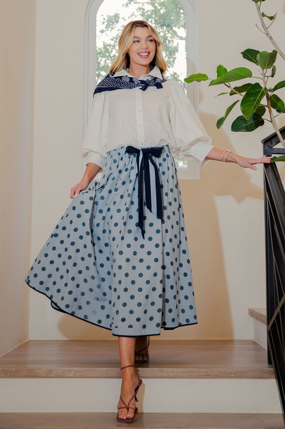 Woman wearing a white blouse and blue polka dot skirt standing indoors.