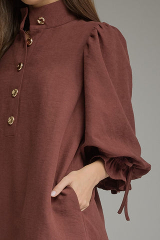 A close-up of a Entro model wearing a merlot dress with a high collar, gold-tone buttons, and voluminous long sleeves with tie details at the cuffs.