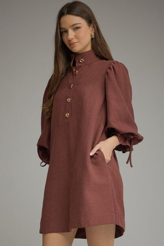 A woman modeling an Entro merlot dress with a high mock neck, gold buttons, and voluminous long sleeves with tie details, set against a solid gray studio background.