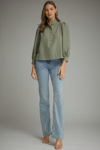 A woman modeling an Entro sage green long-sleeve blouse with a high collar and button placket, paired with light-wash blue jeans and clear high-heel sandals, against a neutral gray studio background.