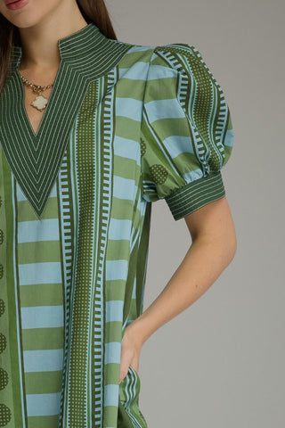 A close-up of a woman wearing a green and light blue patterned short-sleeve top with puff sleeves and a V-neckline, shown against a plain gray studio background.