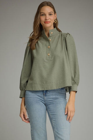 A woman modeling a sage green long-sleeve blouse with a high mock neck, gathered shoulders, and decorative gold buttons, paired with light-wash blue jeans against a solid gray studio background.