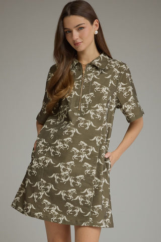 A woman modeling a short-sleeve collared dress with a front zipper and an allover white horse pattern on an olive green base, photographed against a plain gray studio background.