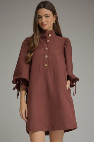 A woman modeling an Entro merlot linen dress with a high mock neck, buttoned chest, puffed elbow-length sleeves with tie cuffs, and side pockets, set against a solid gray studio background.
