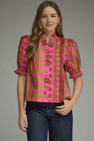 A woman modeling a short-sleeve button-up blouse with a vibrant pink, plum, and olive green patterned design, paired with dark blue jeans.