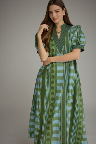 A woman modeling a green and light blue patterned dress with puff sleeves and a V-neckline, photographed against a plain gray studio background.