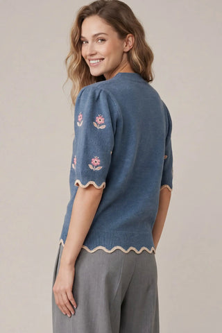 A woman wearing a She & Sky blue short-sleeve knit sweater with pink floral embroidery and scalloped cream trim, paired with gray pants, photographed against a plain beige studio background.