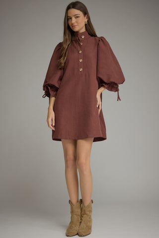 A woman modeling an Entro Merlot puff-sleeve mini dress with a buttoned high collar, styled with tan suede ankle boots against a neutral gray studio background.