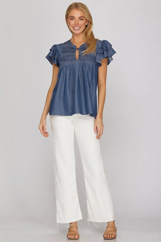 Woman wearing a blue denim She & Sky  blouse with ruffled sleeves and white pants 