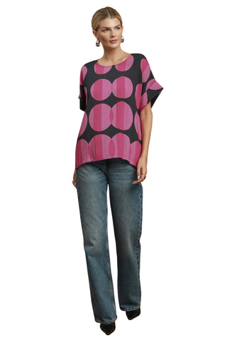 A woman modeling a short-sleeve top with a bold pink and black circular pattern, paired with blue jeans, against a plain white background.