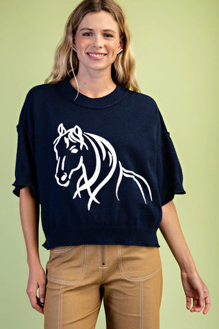 A woman wearing a navy blue crewneck sweater with a white stylized horse graphic on the front, paired with tan pants, set against a solid light green background.