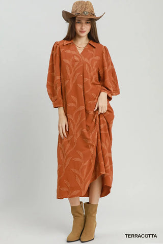 A woman modeling a terracotta-colored midi shirt dress with a tonal leaf pattern, styled with a brown cowboy hat and tan boots against a light gray studio background.