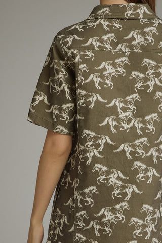 A close-up of a person wearing a short-sleeve button-up shirt with an allover white horse print on an olive green base, shown from the back against a plain gray studio background.