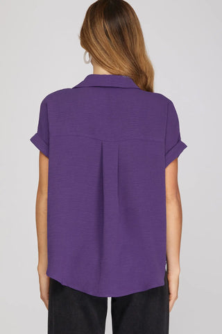 A woman viewed from the back wearing a purple short-sleeve pull on v-neck blouse with a small pleat detail at the center back, paired with black pants.