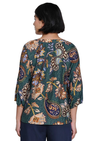 A woman viewed from the back wearing a teal green three-quarter sleeve blouse with a detailed paisley and floral pattern in beige, brown, and blue, paired with navy pants against a white background.
