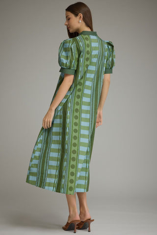 A woman modeling a green and light blue patterned midi dress with puff sleeves, shown from a side-back angle against a neutral gray studio background.