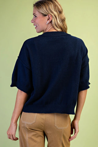 A woman shown from the back wearing a Grigio dark navy blue knit sweater with elbow-length sleeves and tan pants, set against a solid light green background.