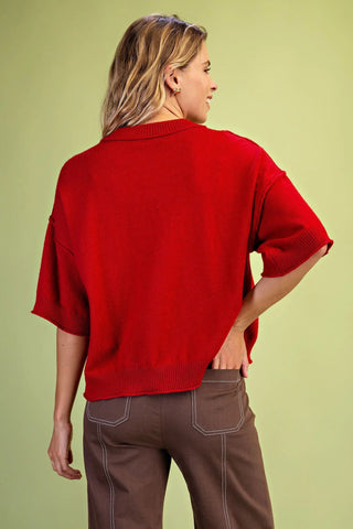 A woman shown from the back wearing a Grigio bright red knit sweater with short wide sleeves and brown pants, standing against a solid light olive-green studio background.