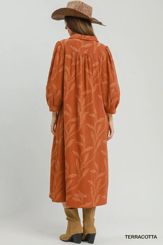 A woman shown from the back wearing a terracotta-colored midi dress by Umgee  with a tonal leaf pattern, paired with tan boots and a brown hat, against a light gray studio background.