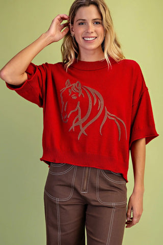A woman wearing a Gigio bright red knit sweater with a large brown horse graphic on the front, paired with brown pants, against a solid light green background.
