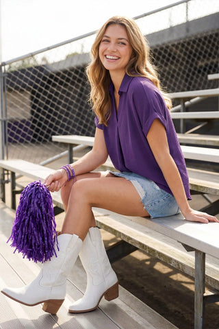 Woman in a She & Sky collared purple shirt and white cowboy boots sitting on bleachers with a purple pom-pom.