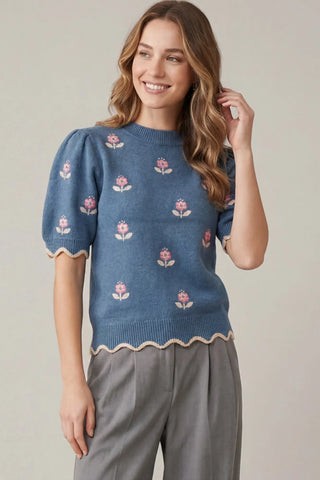 A woman wearing a dusty blue short-sleeve knit sweater with pink and white floral embroidery and scalloped trim, paired with gray trousers, against a neutral beige background.