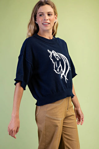 A woman wearing a Gigio navy blue oversized sweatshirt with a white horse graphic on the front, paired with tan pants, against a solid light green background.