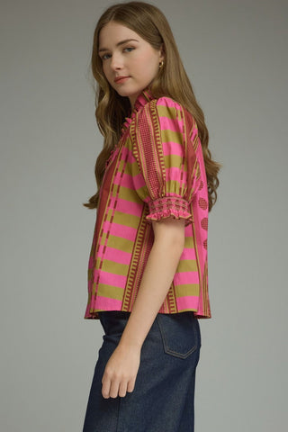 A woman modeling a short-sleeve blouse with pink and olive green horizontal stripes and decorative gold-toned borders, paired with dark blue jeans.