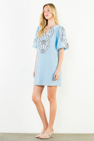 A woman modeling a light blue short dress with silver and white embroidered details on the chest and sleeves, standing against a plain white wall.