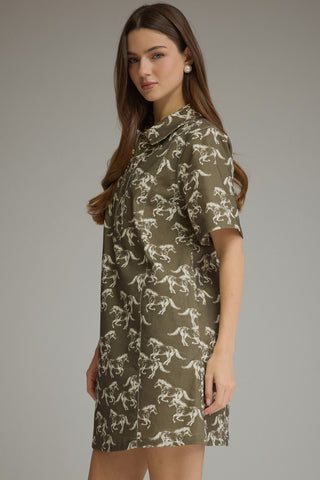 A woman modeling a short-sleeve collared shirt dress with an allover dinosaur print in olive green and off-white, set against a plain gray studio background.