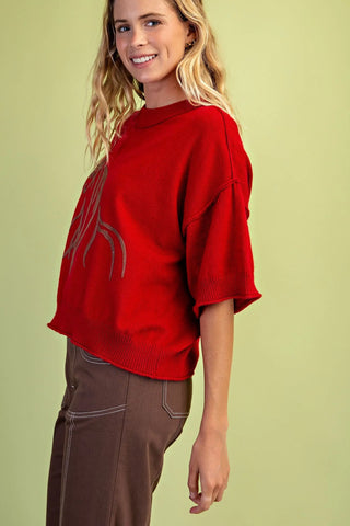 A woman wearing a Grigio bright red knit sweater with a light brown graphic print on the front, paired with brown pants, set against a solid light green background.