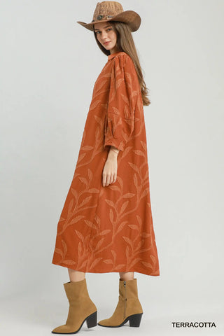 A woman modeling an Umgee terracotta-colored midi dress with a beige leaf pattern, styled with a brown cowboy hat and tan heeled ankle boots against a light gray studio background.