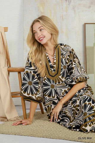 A woman kneeling on a textured rug in a styled indoor setting, wearing a loose, short-sleeve kaftan dress with a bold floral pattern in olive green, black, and cream. The dress features a V-neckline with olive green trim.