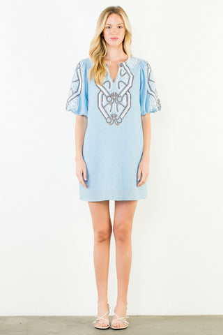 A woman modeling a THML light blue mini dress with short puff sleeves, featuring a V-neckline and a large silver-toned geometric embroidery design on the chest and sleeves.