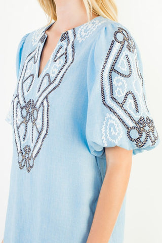 A close-up of a THML light blue linen dress featuring intricate white embroidery and black beaded geometric patterns on the chest and sleeve.