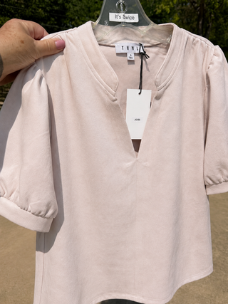 A pale blush pink blouse with a split V-neckline and puff sleeves, held up on a hanger by a hand outdoors. The brand label THML is visible on the collar.